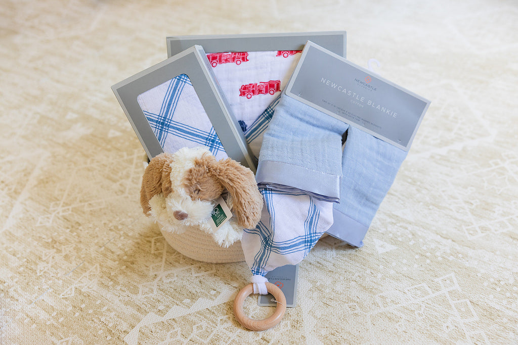 Baby gift set with clothes, a plush toy, and a teething ring on a beige blanket.